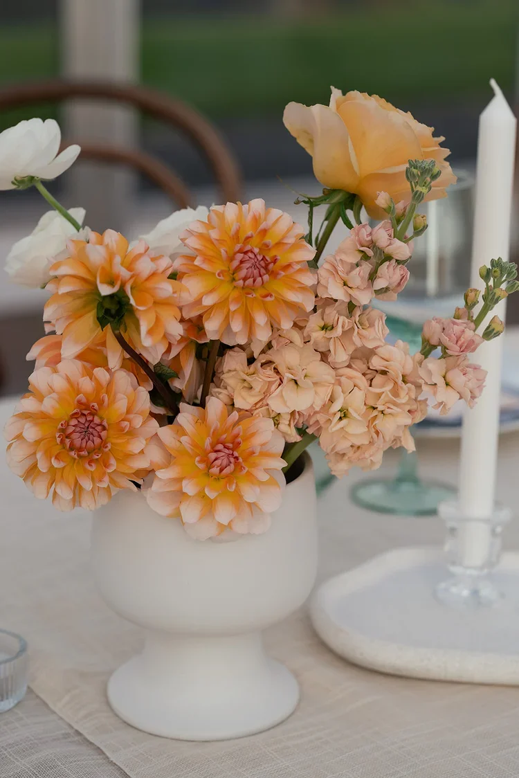 Peach-toned wedding centrepiece featuring dahlias and stock flowers in a modern white vase on a softly textured reception table.