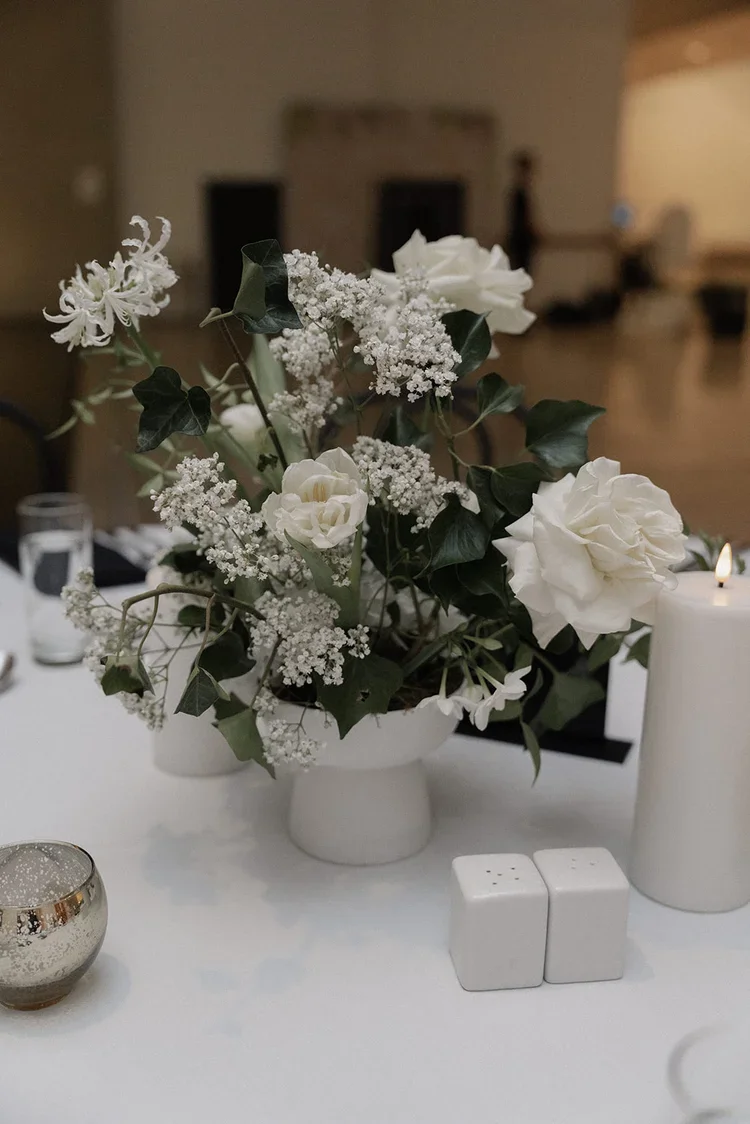 Close-up of an elegant white floral centrepiece featuring roses, baby’s breath, ivy, and textured seasonal blooms on a wedding reception table.