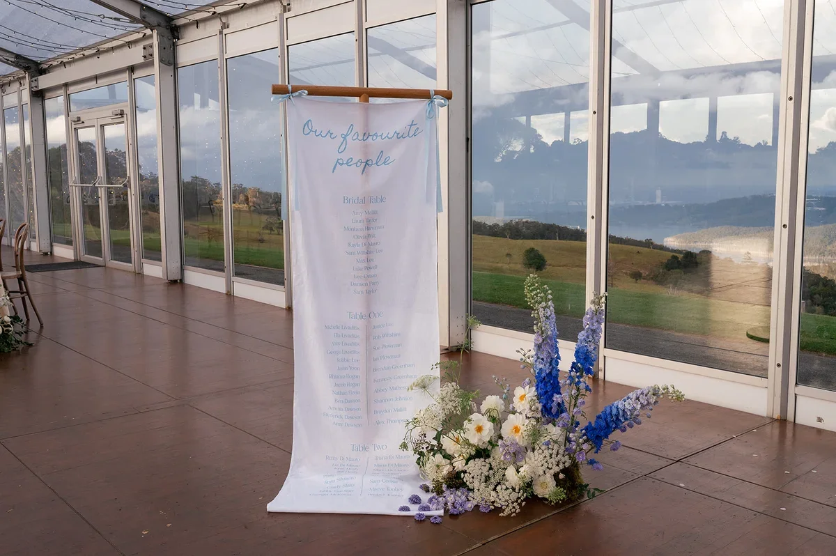 Fabric seating chart displayed beside lush blue and white floral arrangements inside a glass marquee overlooking the Scenic Rim hinterland.”
