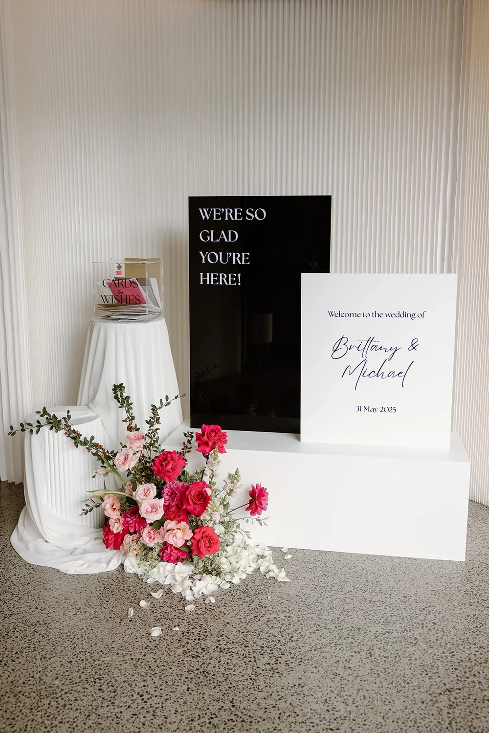 Welcome sign floral centrepiece with roses, ranunculus, and dahlias styled next to plinths and signage. 