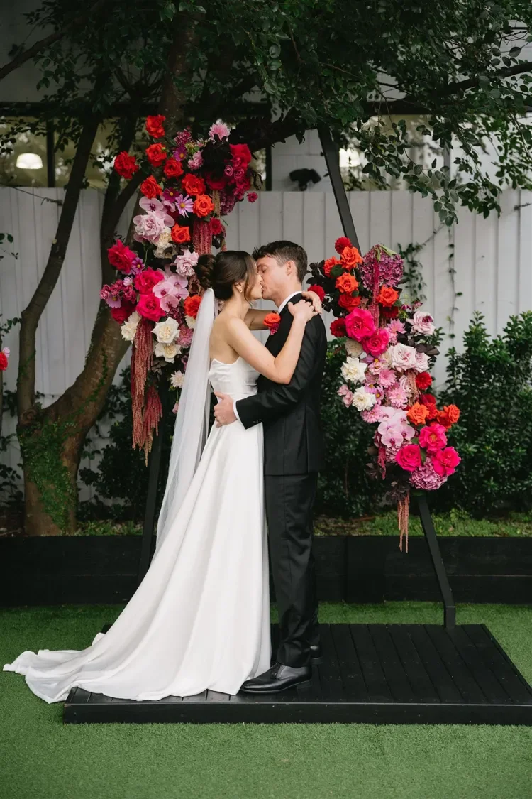 Bride and groom kissing in front of a bold, modern arbour with floral framing them.