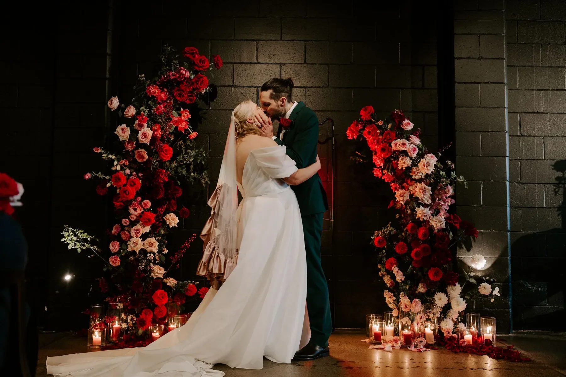 Bride and groom sharing a kiss beneath dramatic red and blush wedding florals and candles at a Brisbane wedding ceremony