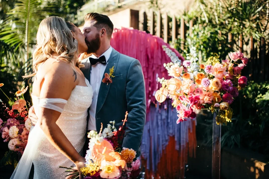 Sarah and Marshall sharing a kiss surrounded by colourful florals and vibrant streamer backdrop during their wedding portraits.