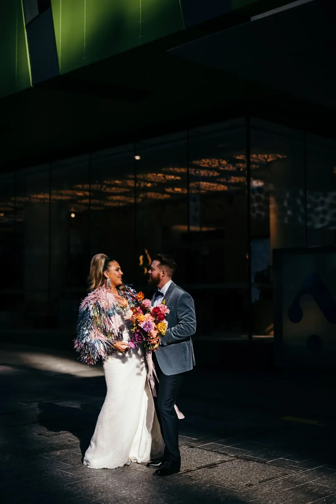 Sarah and Marshall standing together in dramatic light, holding their vibrant wedding bouquet in an urban city setting.