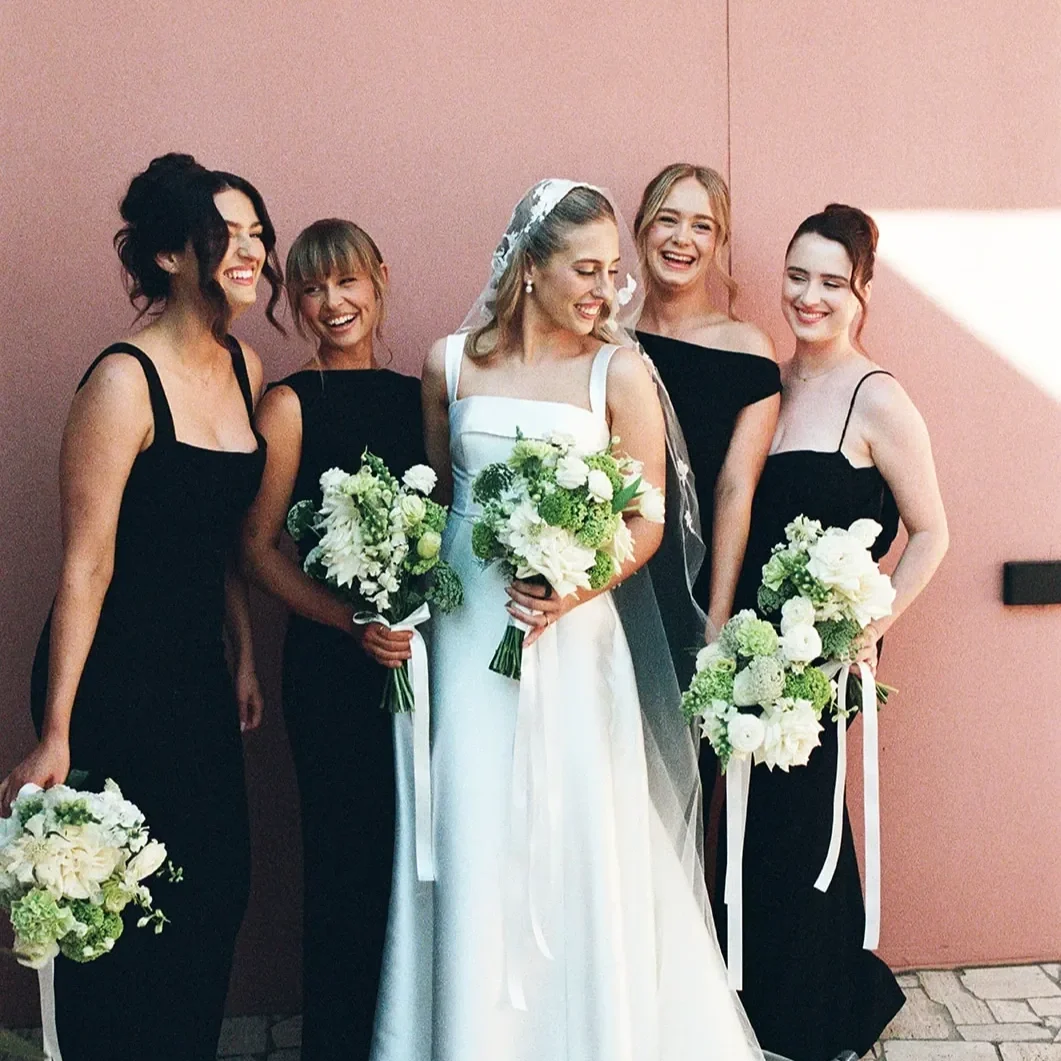 Bride and bridesmaids holding white and green bouquets against a soft pink wall, florals by a Brisbane wedding florist.