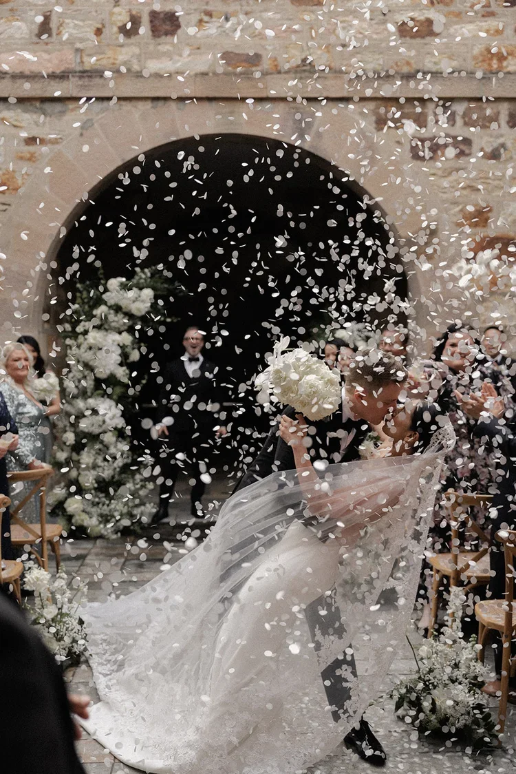 Bride and groom kissing under a dramatic shower of white confetti during their ceremony exit.