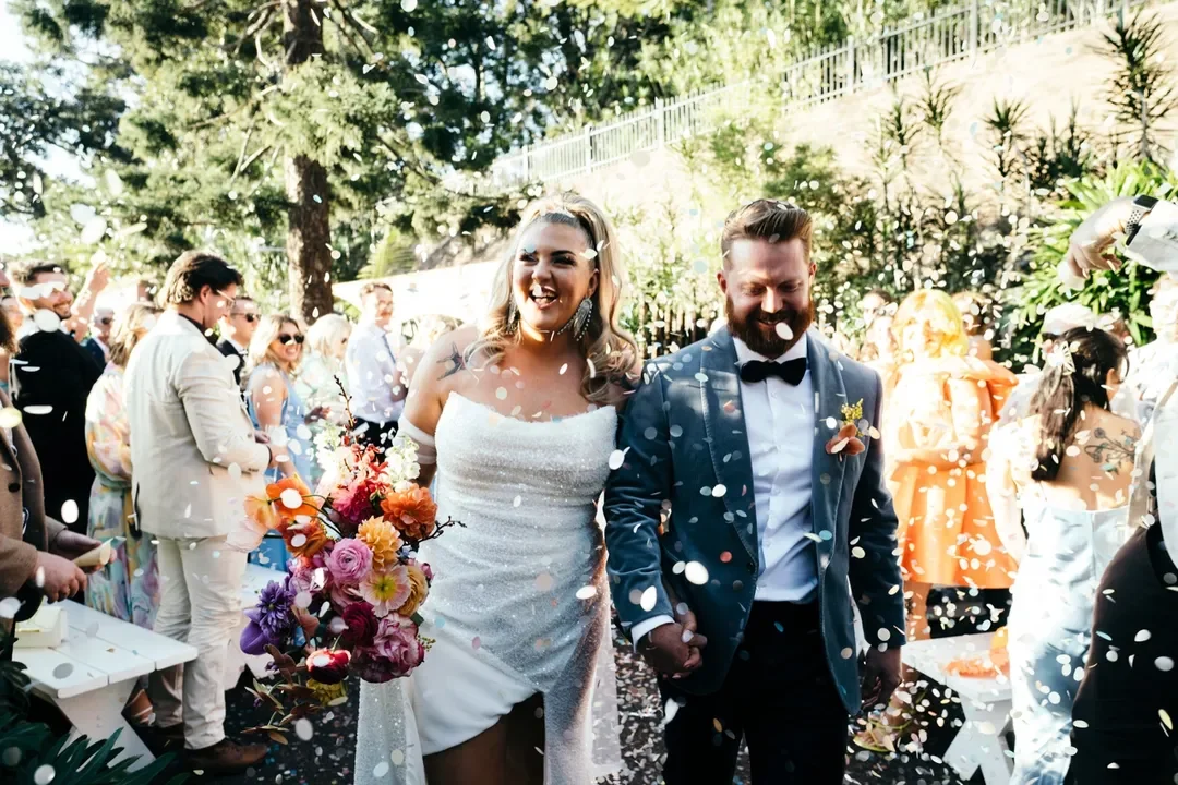 Sarah and Marshall walking through a shower of confetti as wedding guests celebrate around them outdoors.