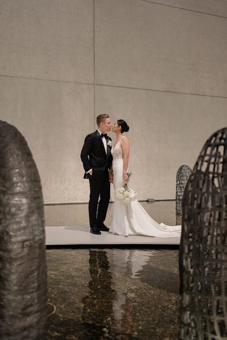 Bride and groom standing together on a platform above reflective water inside the Gallery of Modern Art, holding hands and smiling softly at each other.