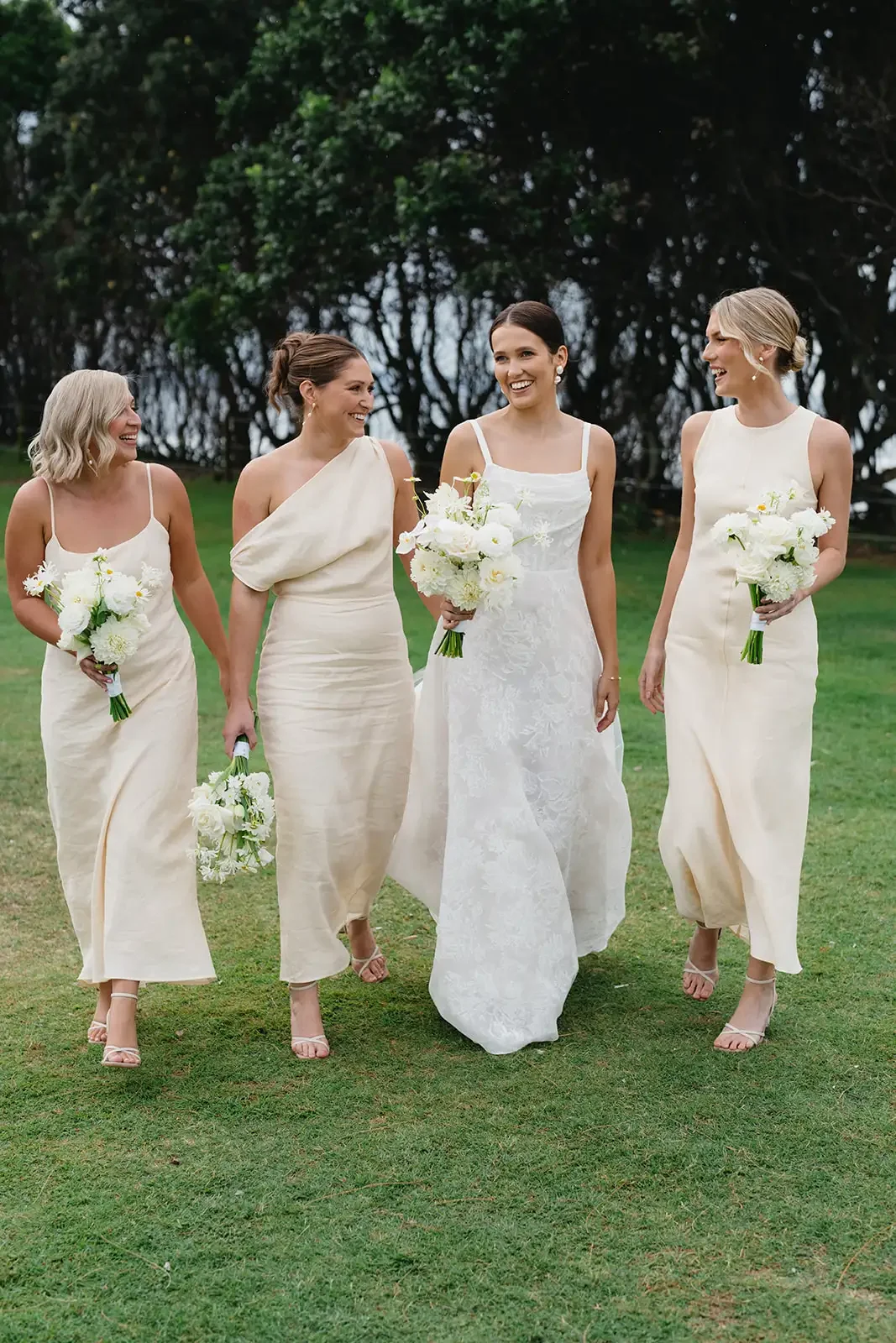 Bride and bridesmaids in neutral dresses walking with white wedding bouquets on the lawn.