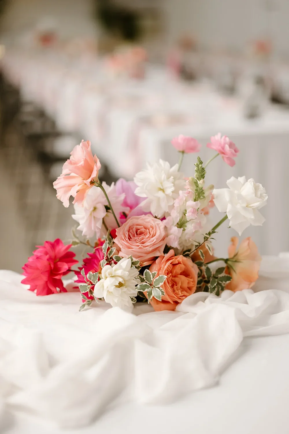 Pastel floral centrepiece with peach roses, pink ranunculus, and white dahlias styled on a draped white tablecloth.