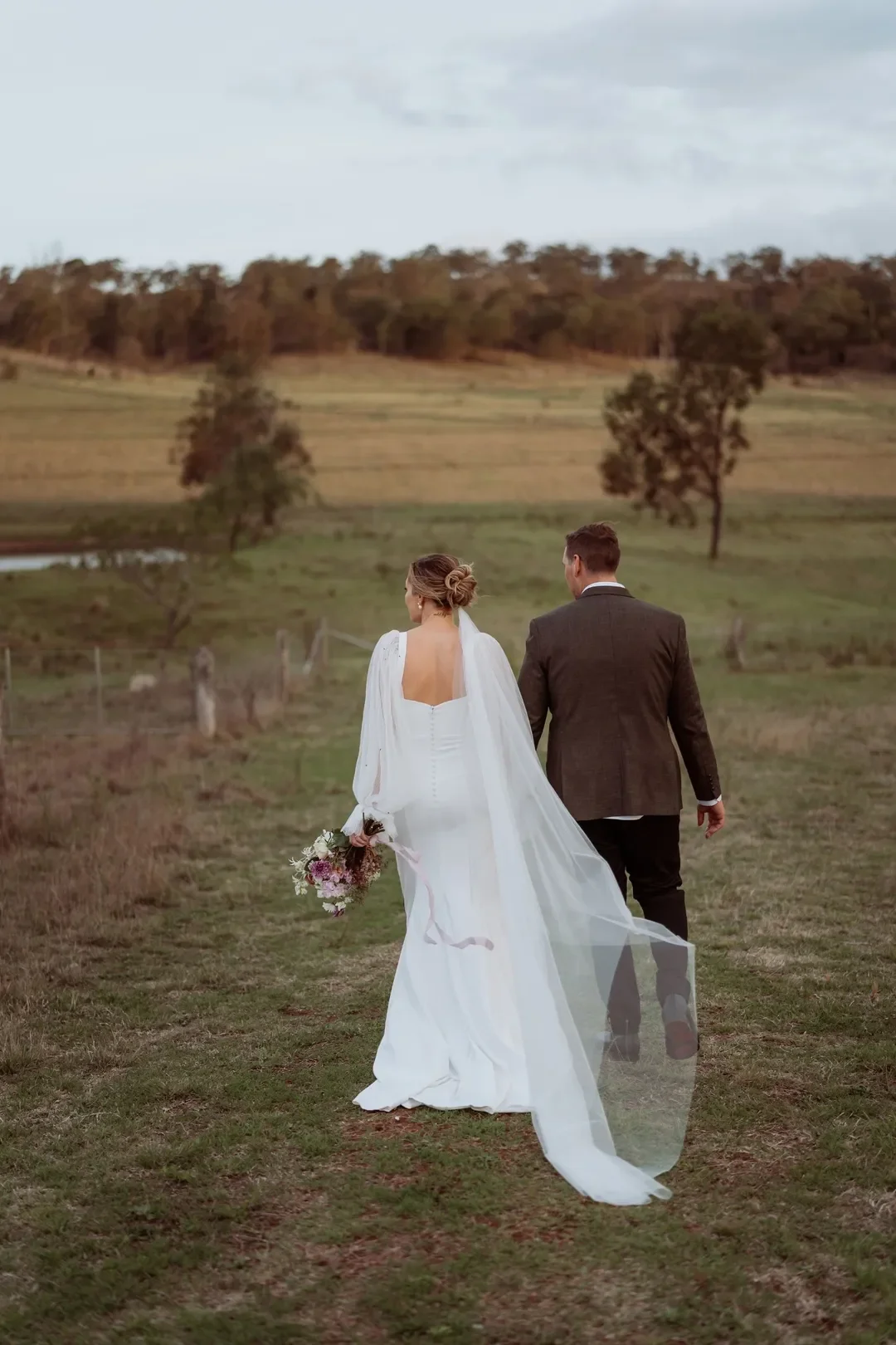 Bride and groom walking hand-in-hand across open countryside at sunset, with Annalise’s veil trailing behind her and bouquet in hand.
