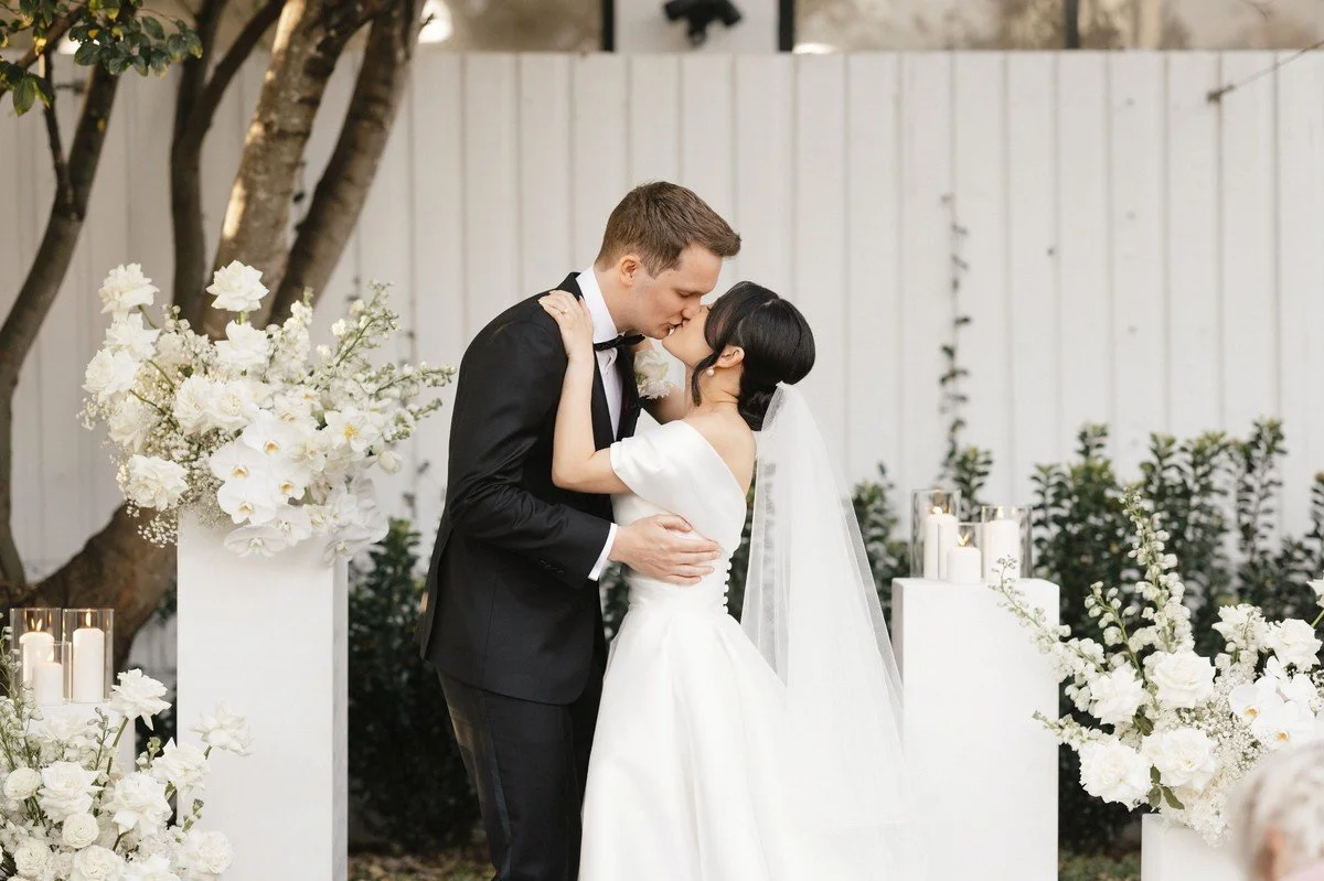 Bride and groom sharing their first kiss surrounded by modern white floral arrangements and tall pillar stands.