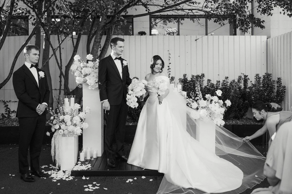 Bride and groom standing at the ceremony altar while a bridesmaid adjusts the bride’s veil among white floral pillars.