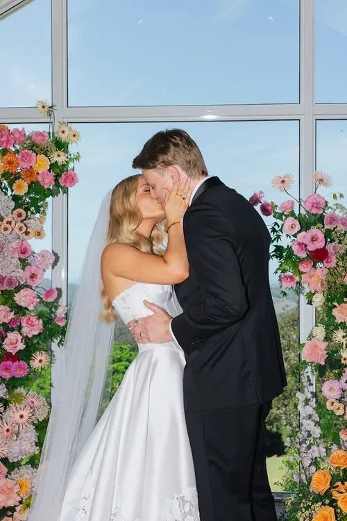 Bride and groom sharing a kiss between vibrant floral ceremony pillars by Garden Graffiti