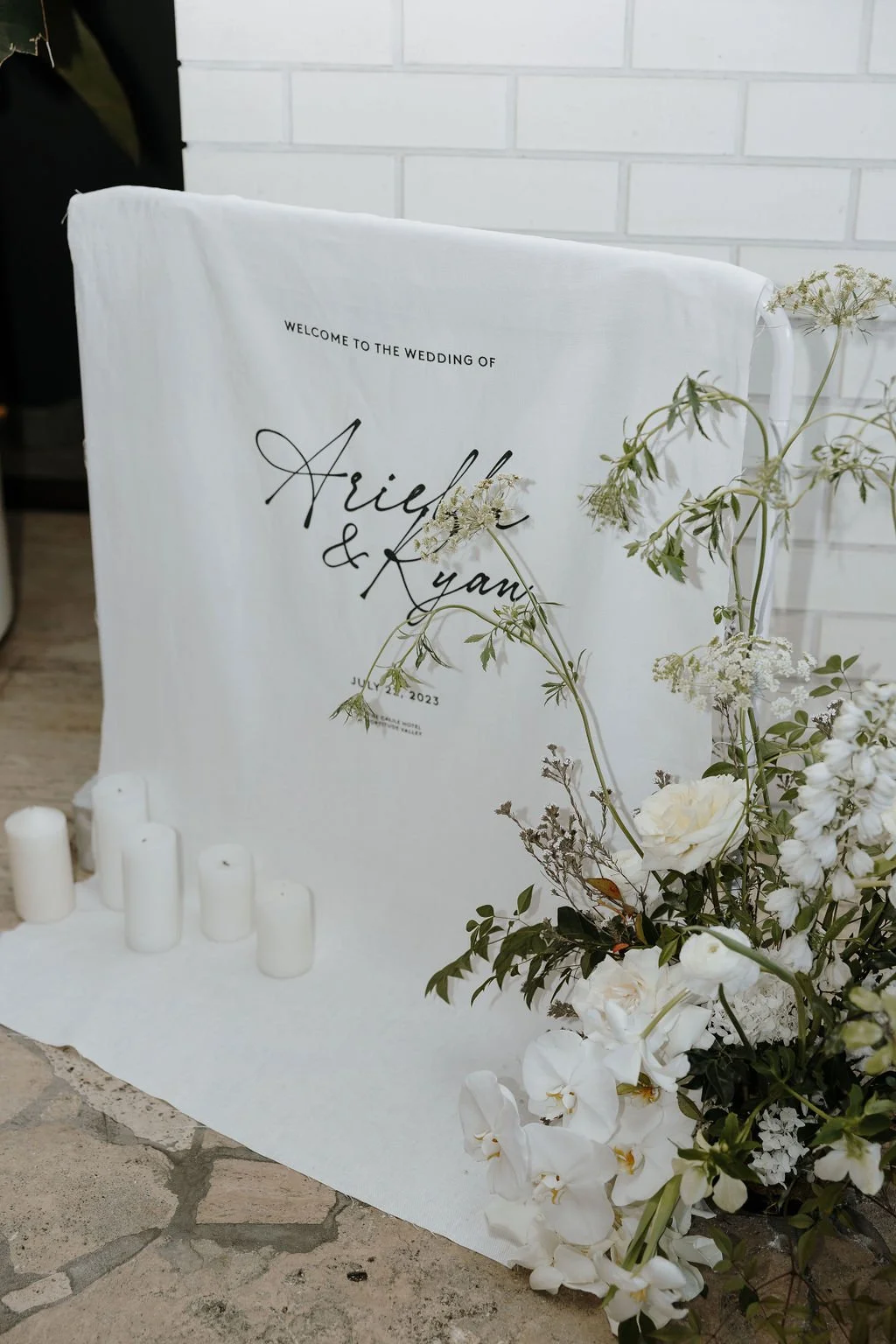 Wedding welcome sign draped in fabric with surrounding white florals and pillar candles.