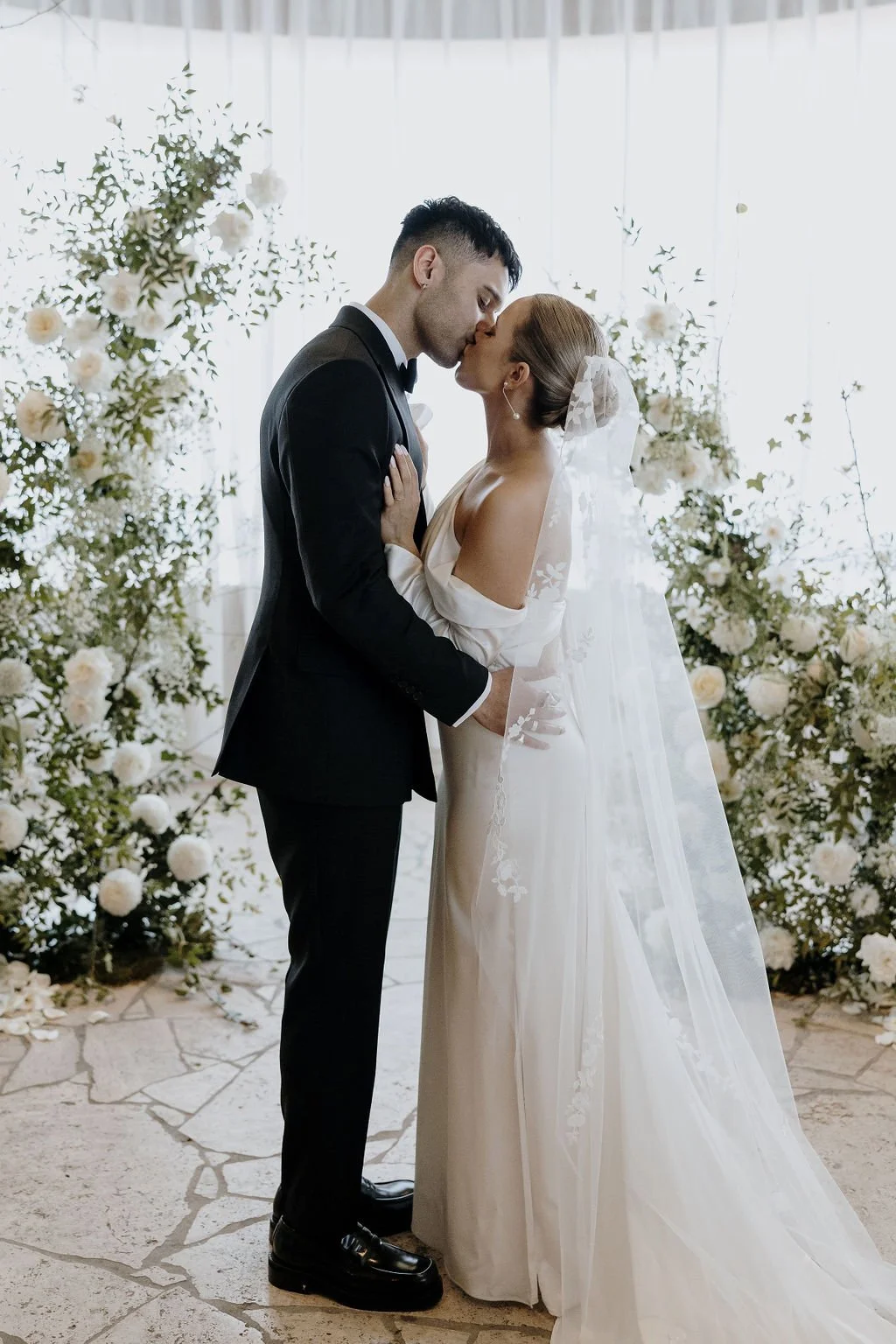 Bride and groom sharing a kiss amid lush white floral pillars at their ceremony.