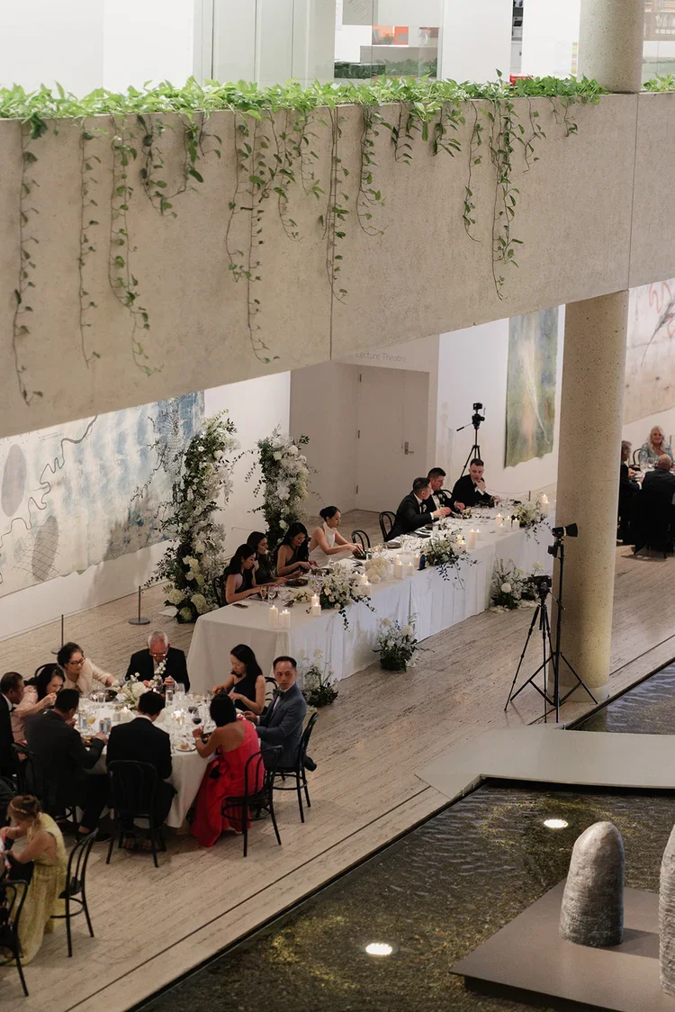 Overhead view of Vy & Sean’s wedding reception at GOMA, showing the long bridal table, floor florals, and guests dining beside the water feature.