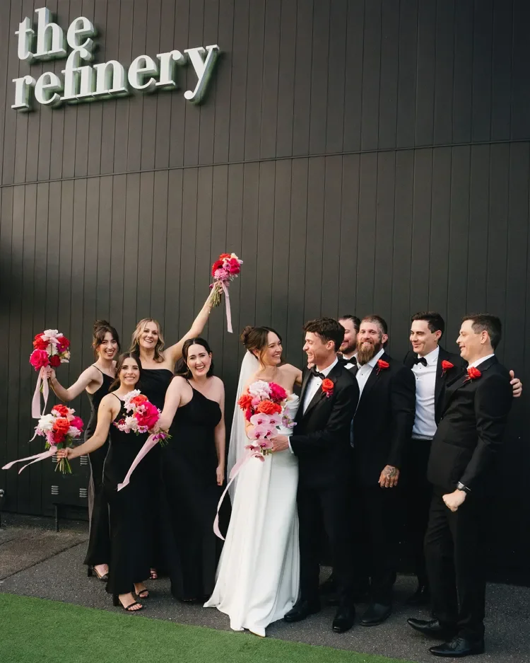 Bridal party cheering together outside The Refinery, holding bright bouquets and dressed in mixed tones.