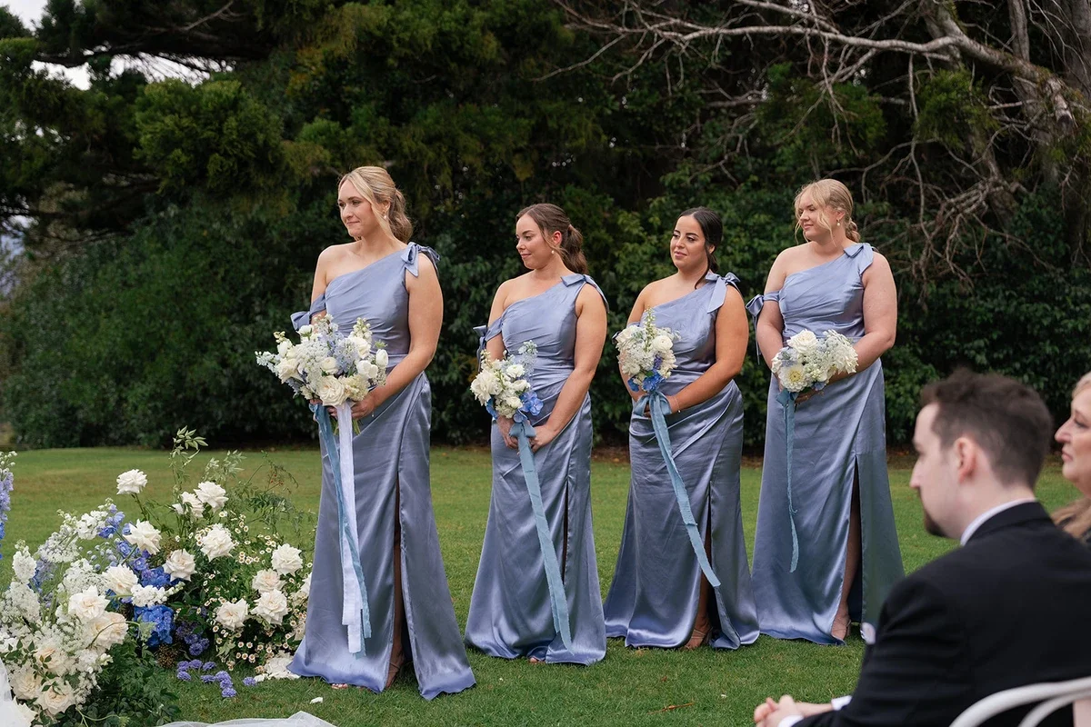 Bridesmaids in soft blue satin gowns holding white and blue bouquets during the outdoor wedding ceremony.”