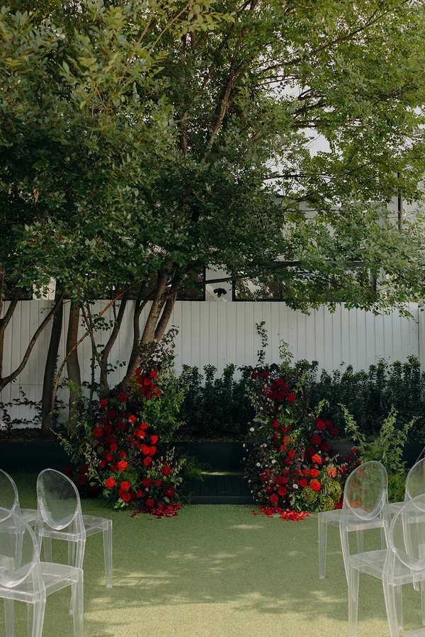 Outdoor ceremony space styled with vibrant red and orange florals and transparent ghost chairs.