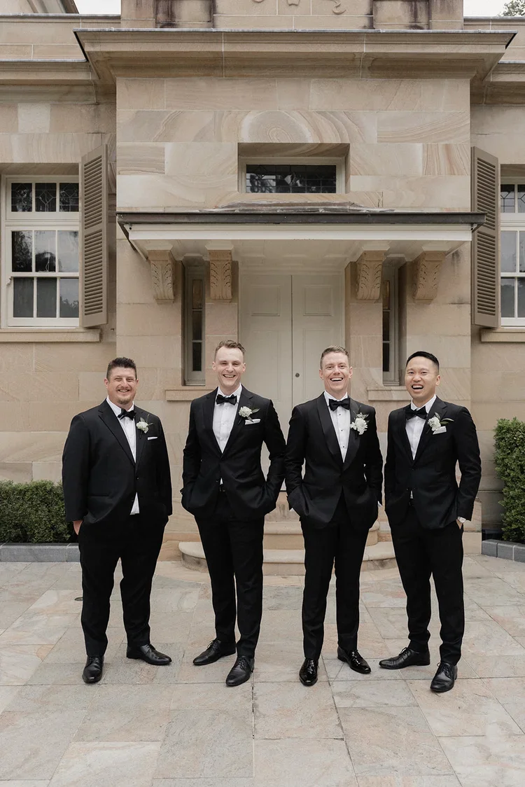 Groom with his groomsmen standing in front of a heritage sandstone building in classic black suits and bow ties.
