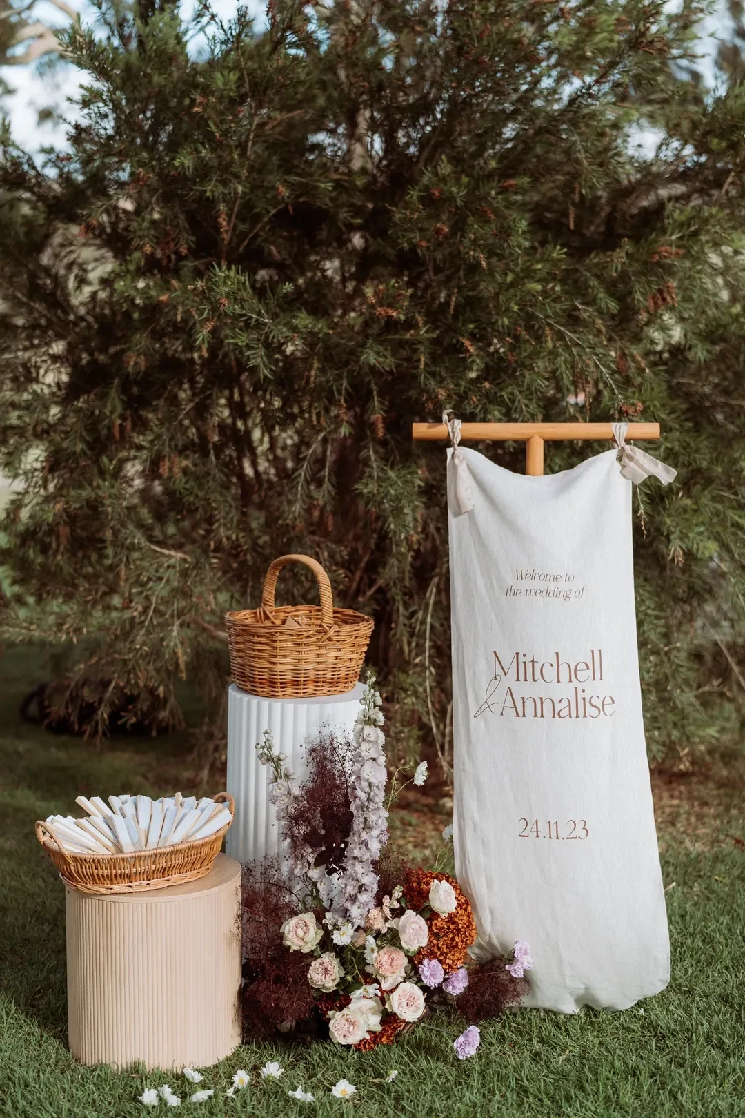 Wedding welcome display with personalised signage, baskets, and floral arrangements against a natural backdrop.