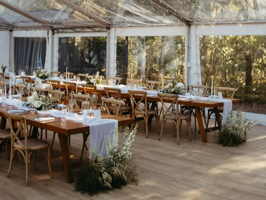 Clear marquee wedding reception with natural timber tables, soft white floral arrangements, and airy forest views