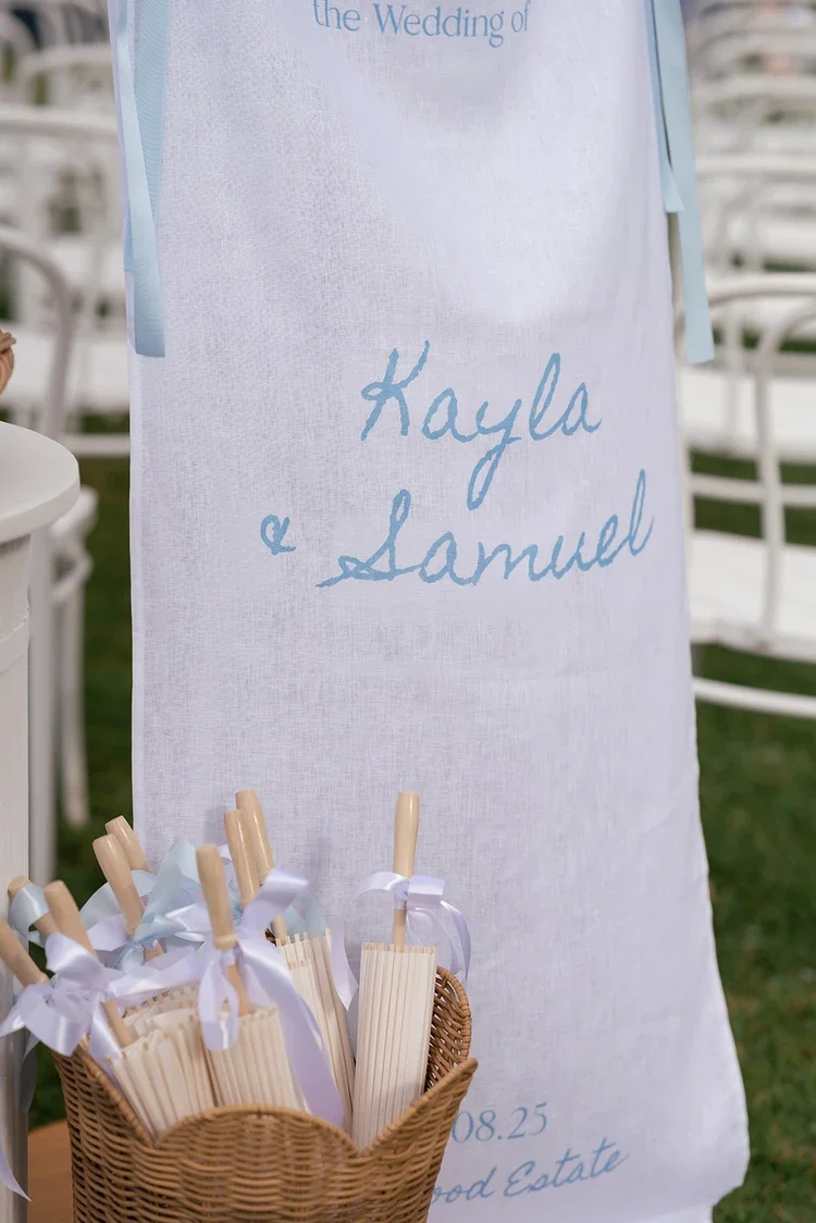 Wedding welcome banner with Kayla and Samuel’s names displayed beside a basket of guest parasol fans tied with pastel ribbons.