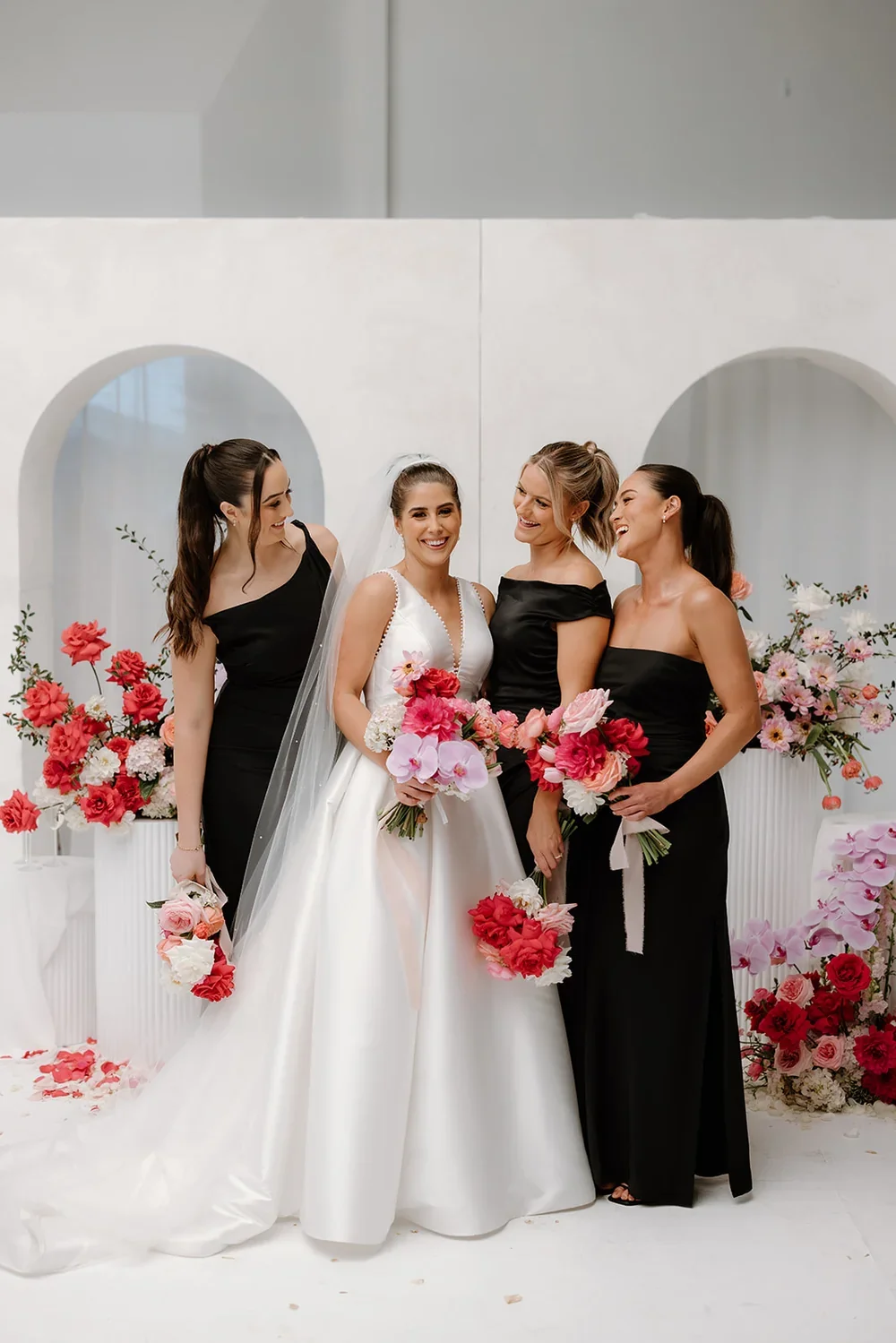 White bridal party bouquets with soft peach, blush, and lavender florals held by bridesmaids in black dresses.