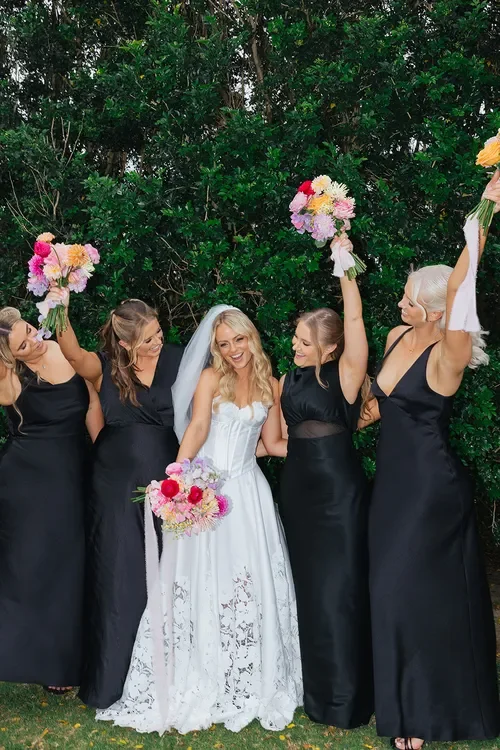 Bride celebrating with her bridesmaids holding colourful Garden Graffiti bouquets, wearing black dresses outdoors after the ceremony