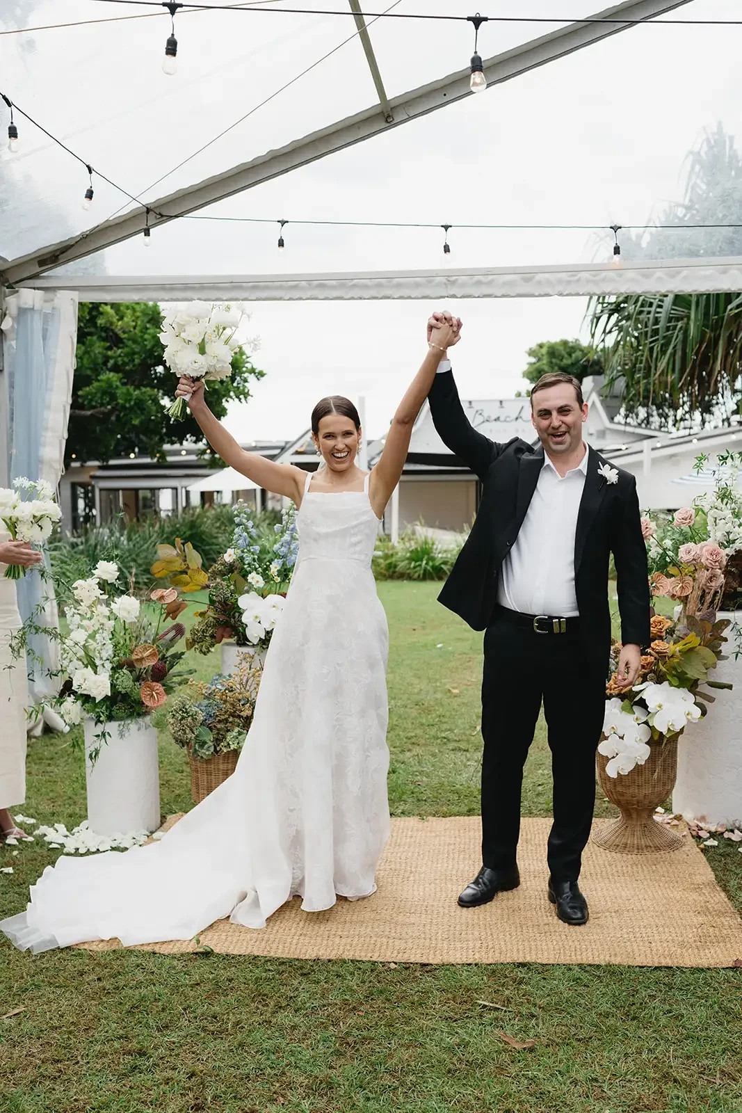 Bride and groom celebrating under a clear marquee with lush garden wedding flowers in Byron Bay.