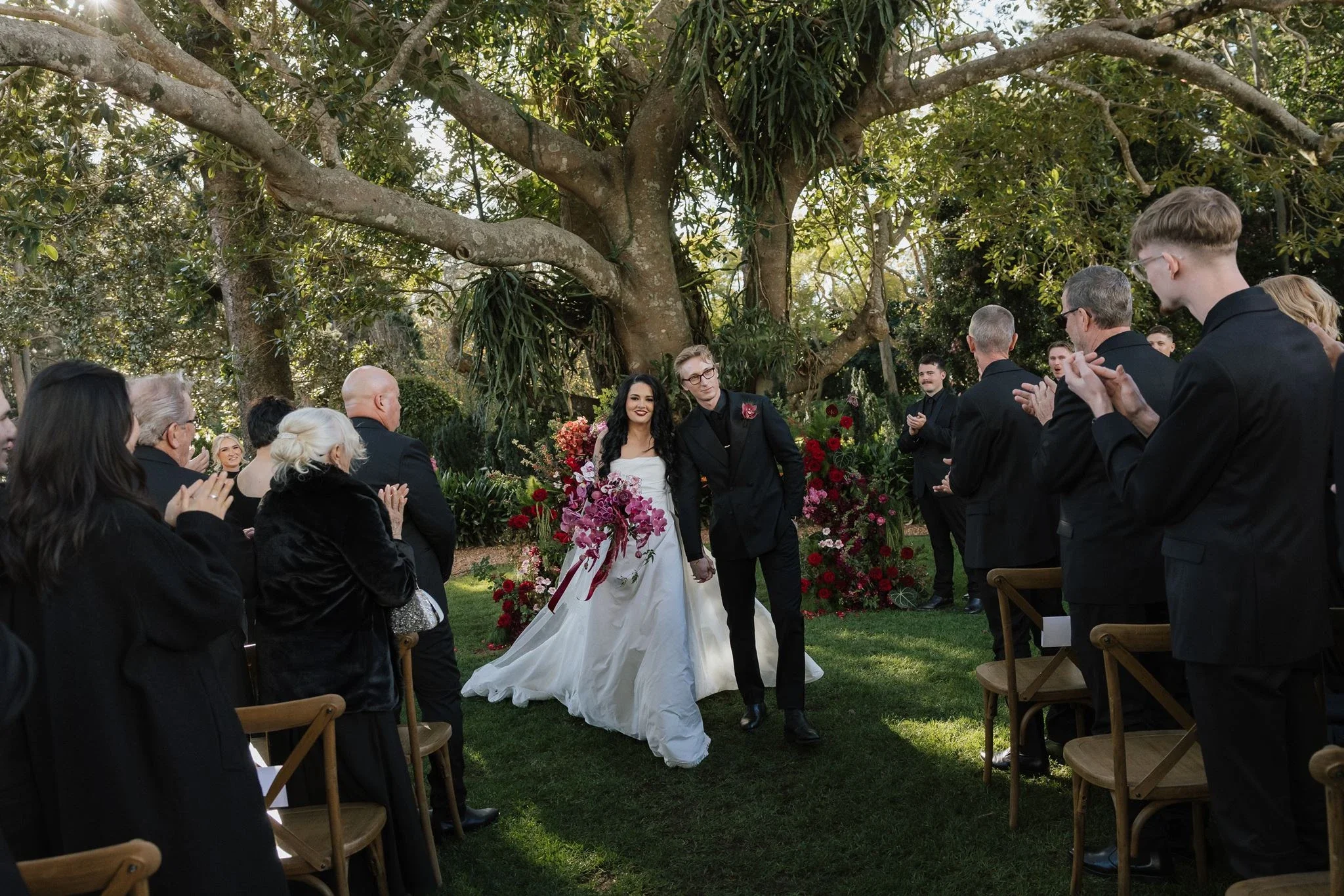 Bride and groom walking back up the aisle past guests dressed in black under the trees at Gabbinbar Homestead