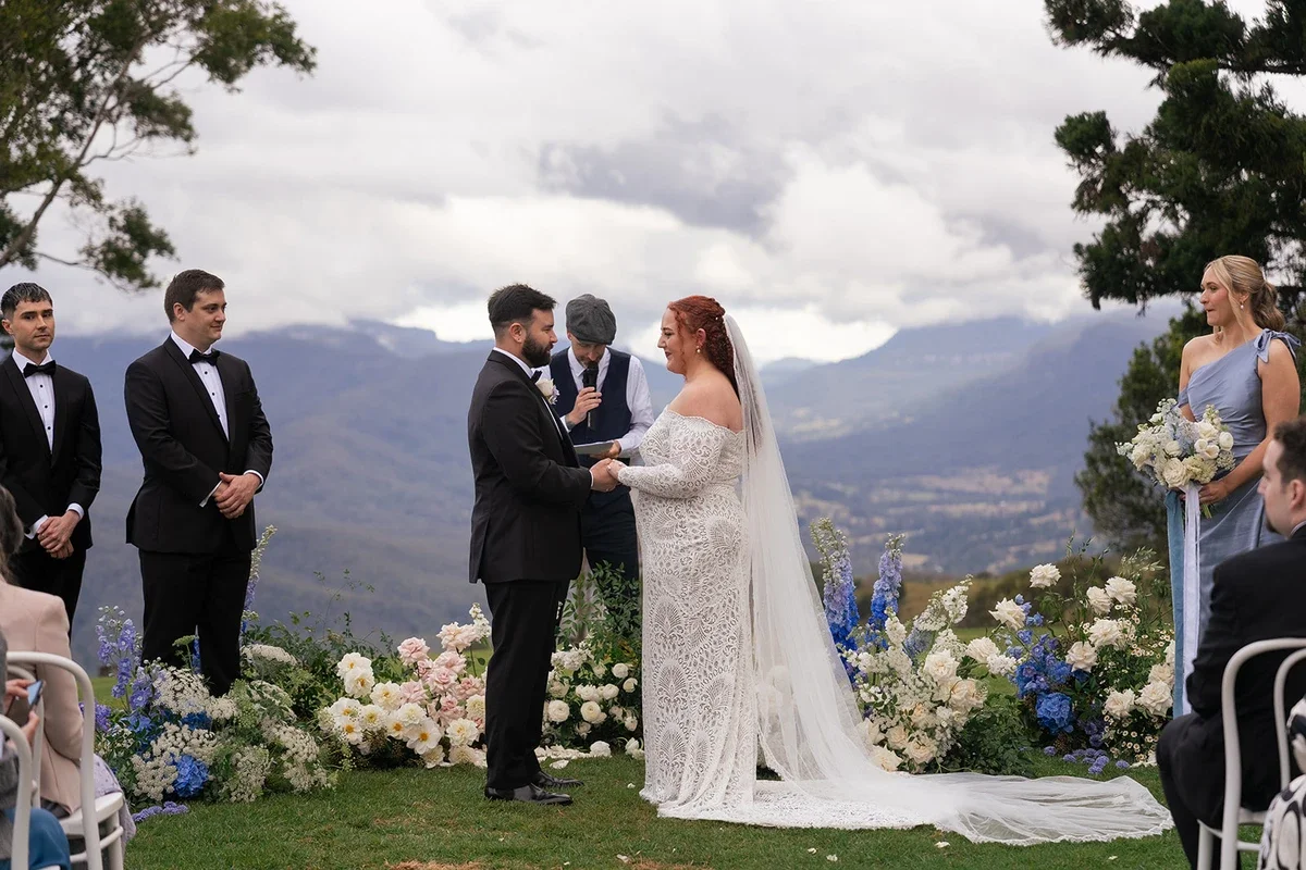 Outdoor wedding ceremony with panoramic mountain views, blue and white floral arrangements, and the couple holding hands during vows.