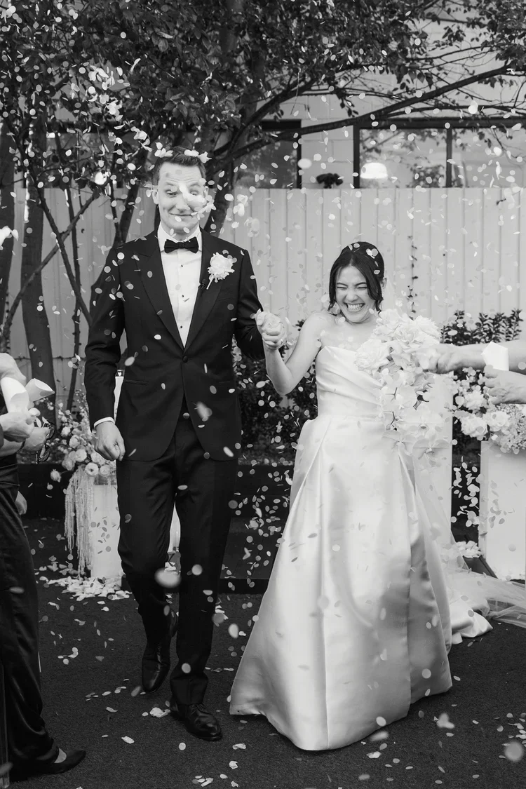 Bride and groom walking through a joyful petal toss, smiling as flower petals fall around them.