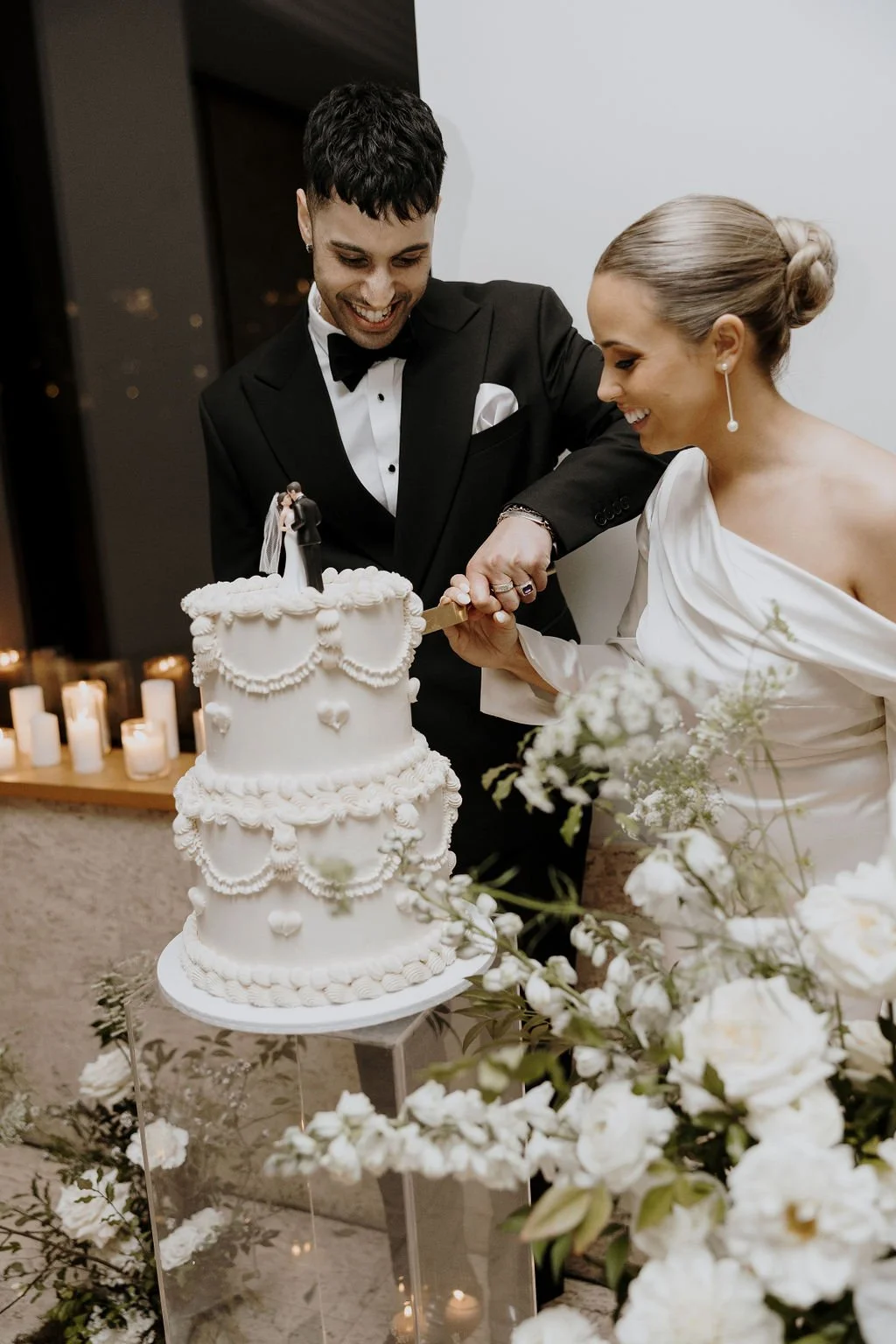 Bride and groom cutting their vintage-inspired white wedding cake surrounded by white florals.
