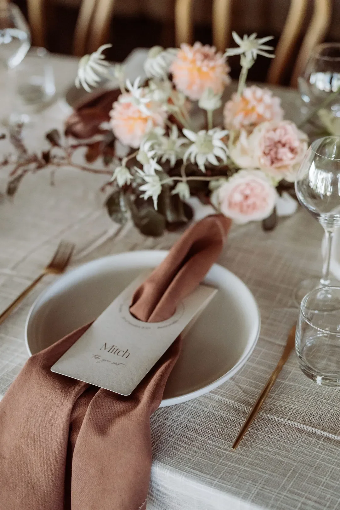 Wedding place setting with soft brown napkin, personalised name card, and textured blush floral centrepiece.