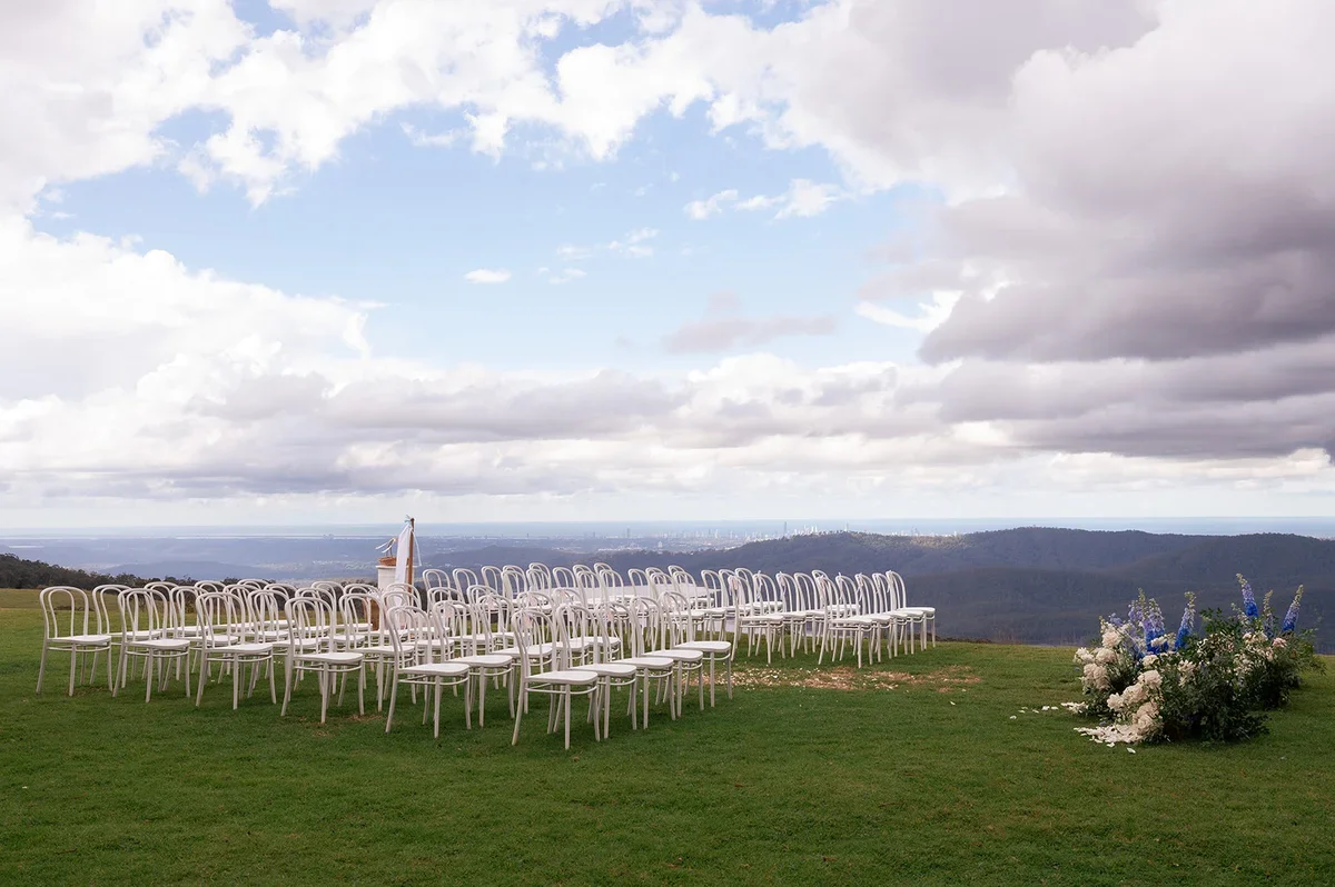 Wide landscape view of Kayla and Sam’s mountain-top wedding ceremony setup with white chairs, blue and white floral arrangements, and sweeping hinterland scenery.