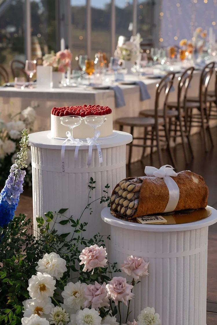 Wedding cake and giant cannoli dessert display on white pedestal stands, surrounded by soft pastel florals at a Scenic Rim marquee reception.