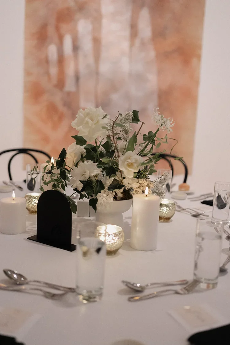 Wedding reception table decorated with white flowers, pillar candles, small gold votives, and modern black table numbers.