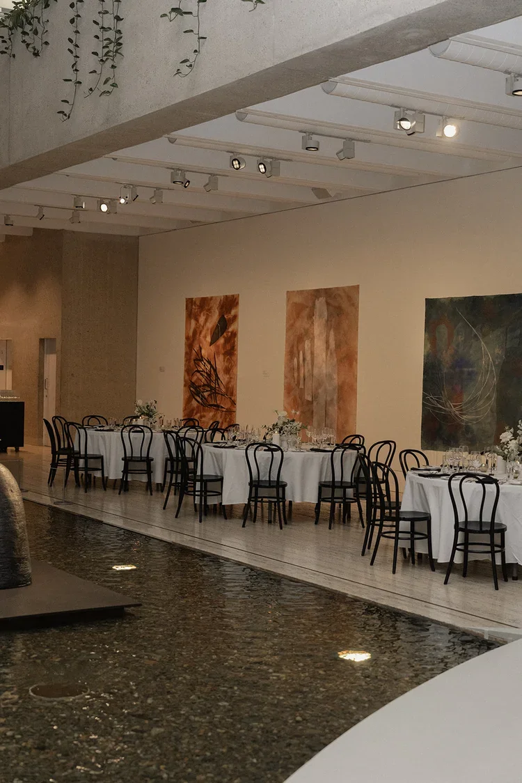 Wide shot of minimalist wedding reception tables at GOMA with white linens, black chairs, modern art on the walls, and a reflecting pool beside the space.
