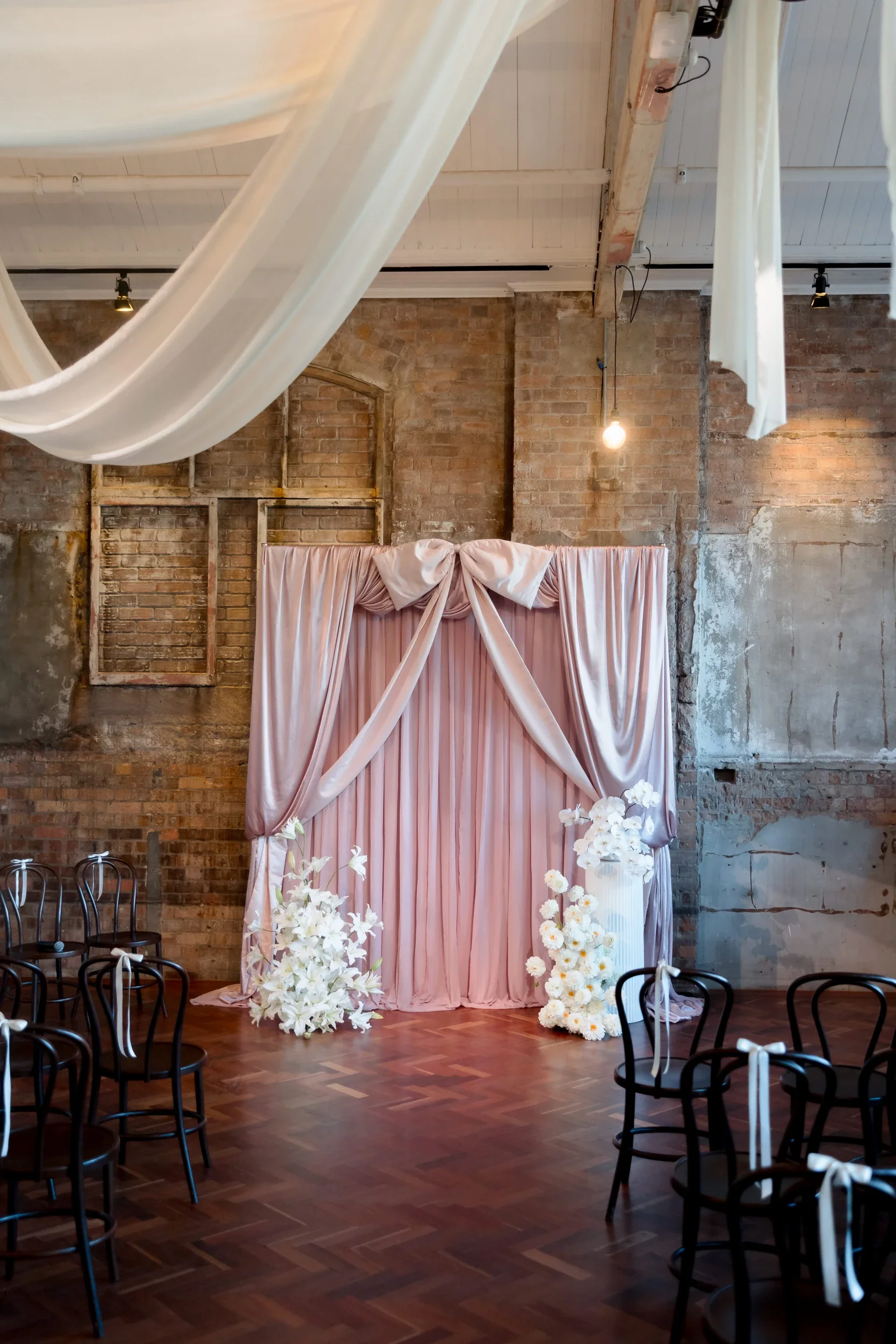 Front-on view of a blush draped ceremony space with white floral columns and ceiling swags at The Warehouse Brisbane