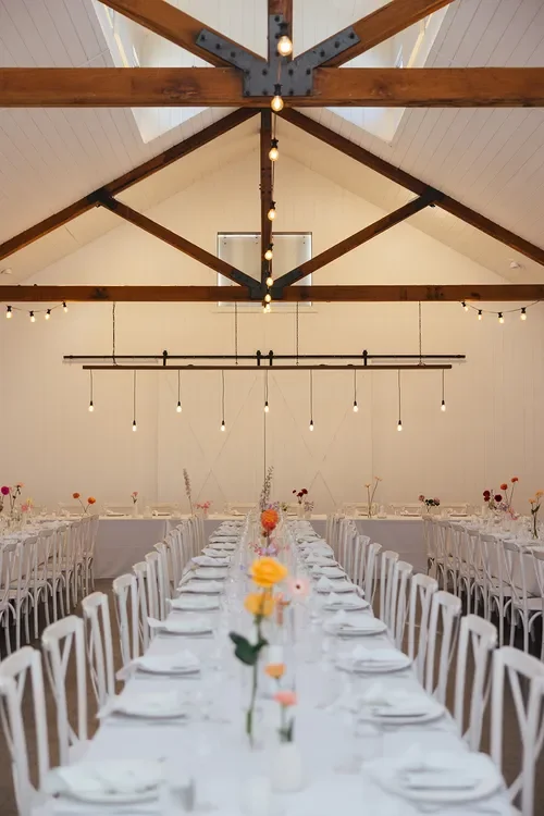 Long wedding reception table styled with delicate Garden Graffiti bud vase florals beneath timber beams and soft lighting