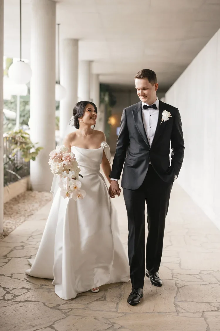 Bride and groom walking hand-in-hand under the Calile Hotel walkway, with the bride holding a soft blush and white orchid bouquet.
