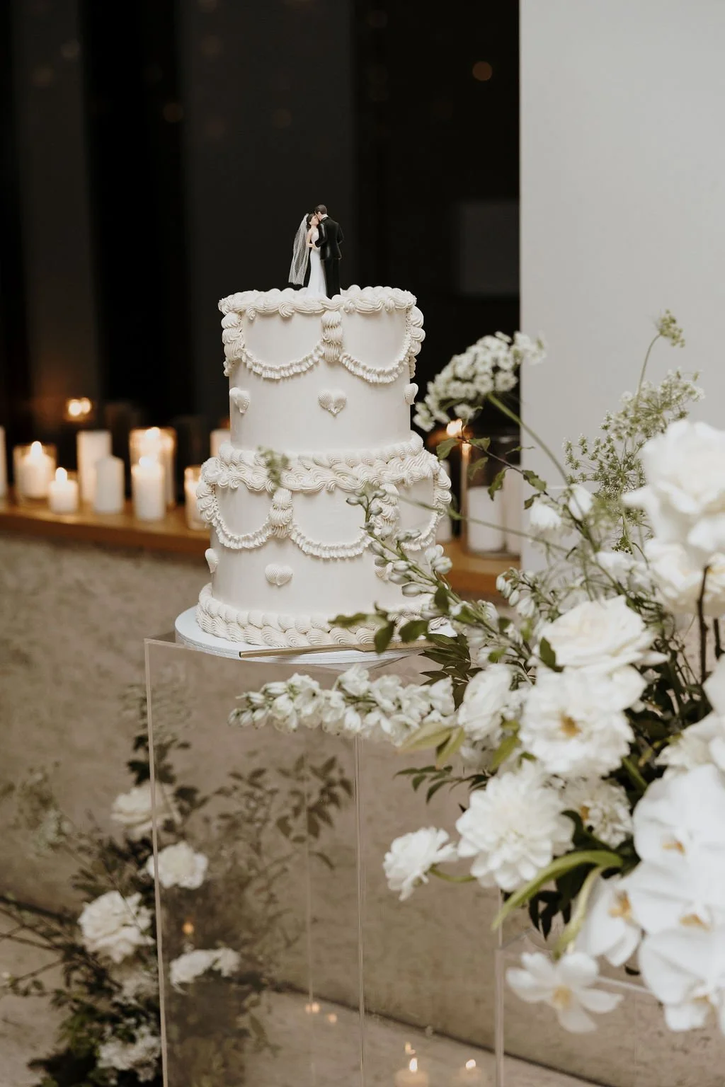 Three-tier vintage-inspired white wedding cake with piped details surrounded by soft white florals.