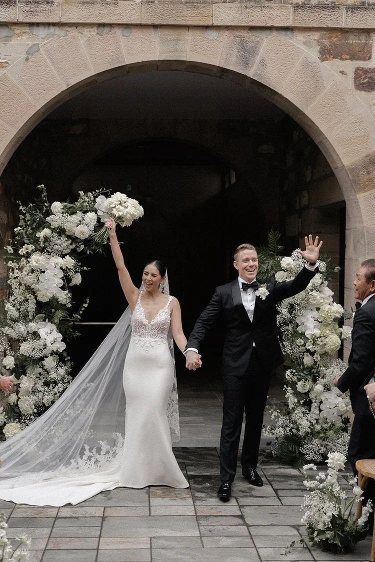 Bride and groom celebrating joyfully while walking back down the aisle, raising their hands and bouquet in excitement.
