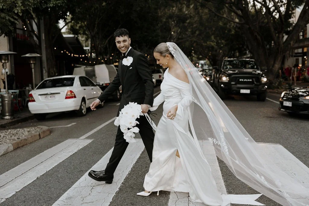Bride and groom walking across James Street, veil flowing and carrying cascading orchid bouquet.