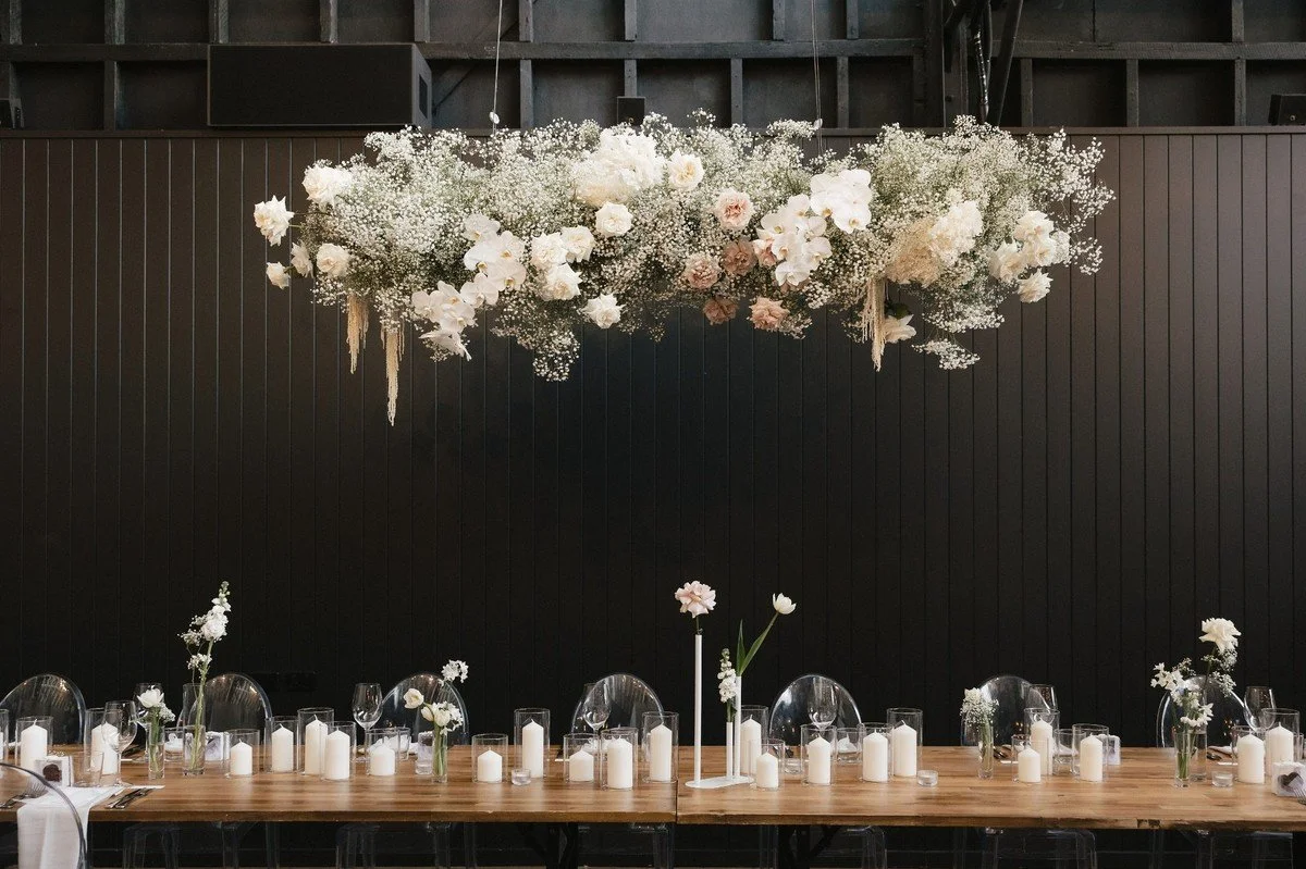 Elegant long reception table with ghost chairs, candles, and single-stem white florals under a hanging cloud installation.