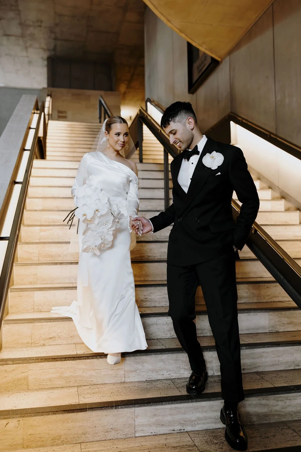 Bride and groom walking down modern stone staircase, bride holding cascading orchid bouquet.