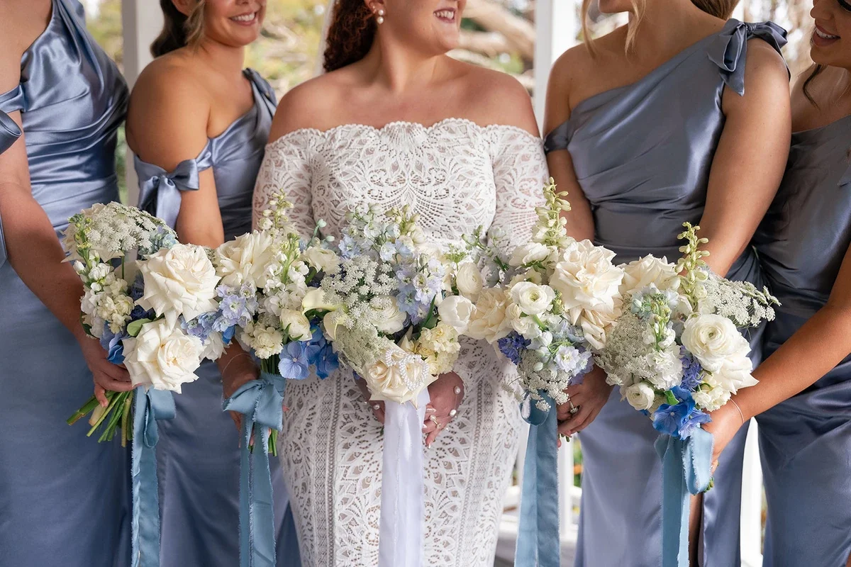 Bride and bridesmaids holding soft blue and white bouquets with silk ribbons, featuring roses, delphiniums, and Queen Anne’s lace.