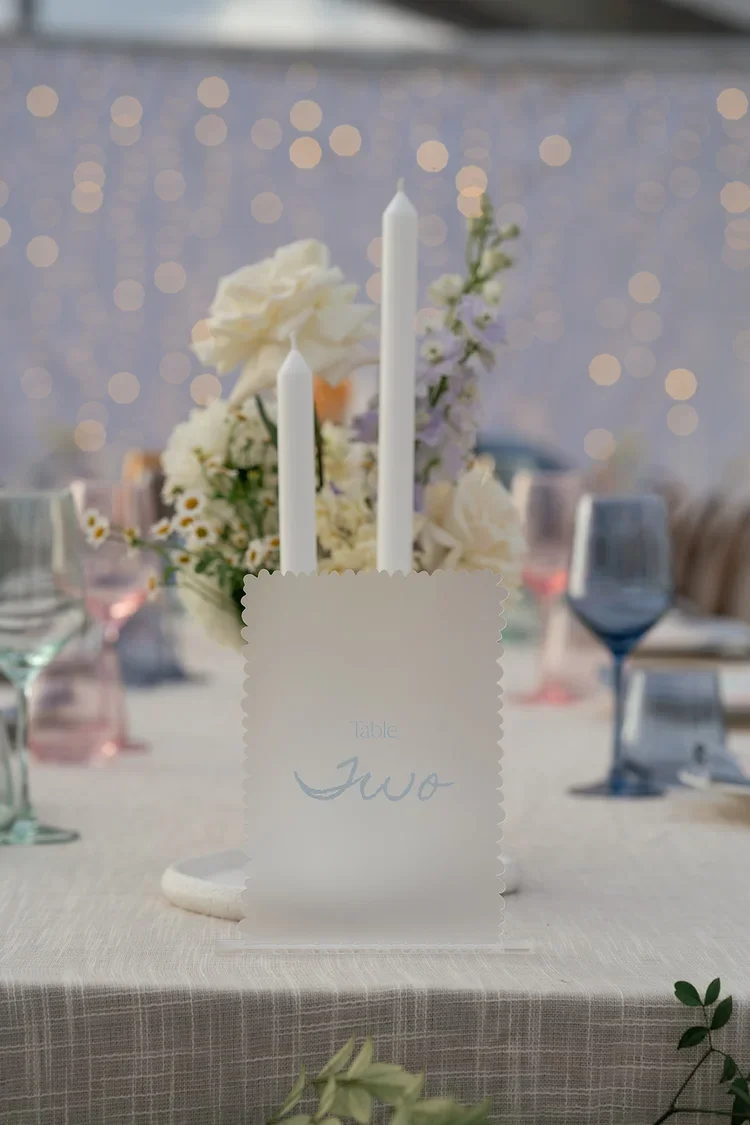 Close-up of Kayla and Sam’s wedding table décor featuring pastel flowers, white taper candles and a frosted acrylic ‘Table Two’ sign with twinkling lights in the background.”